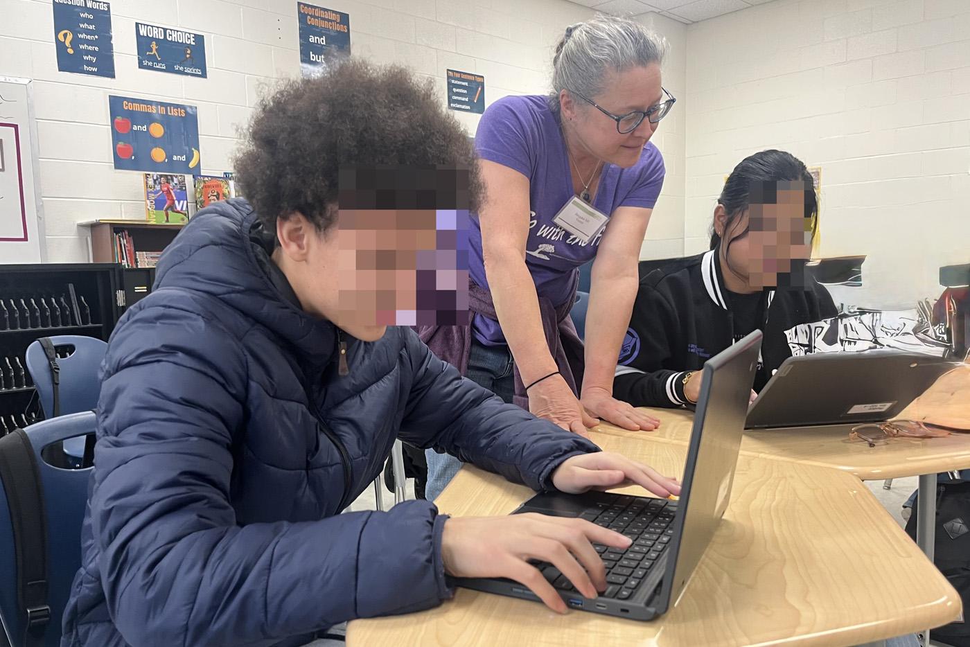 Female teacher leans over to help two students on laptop computers.