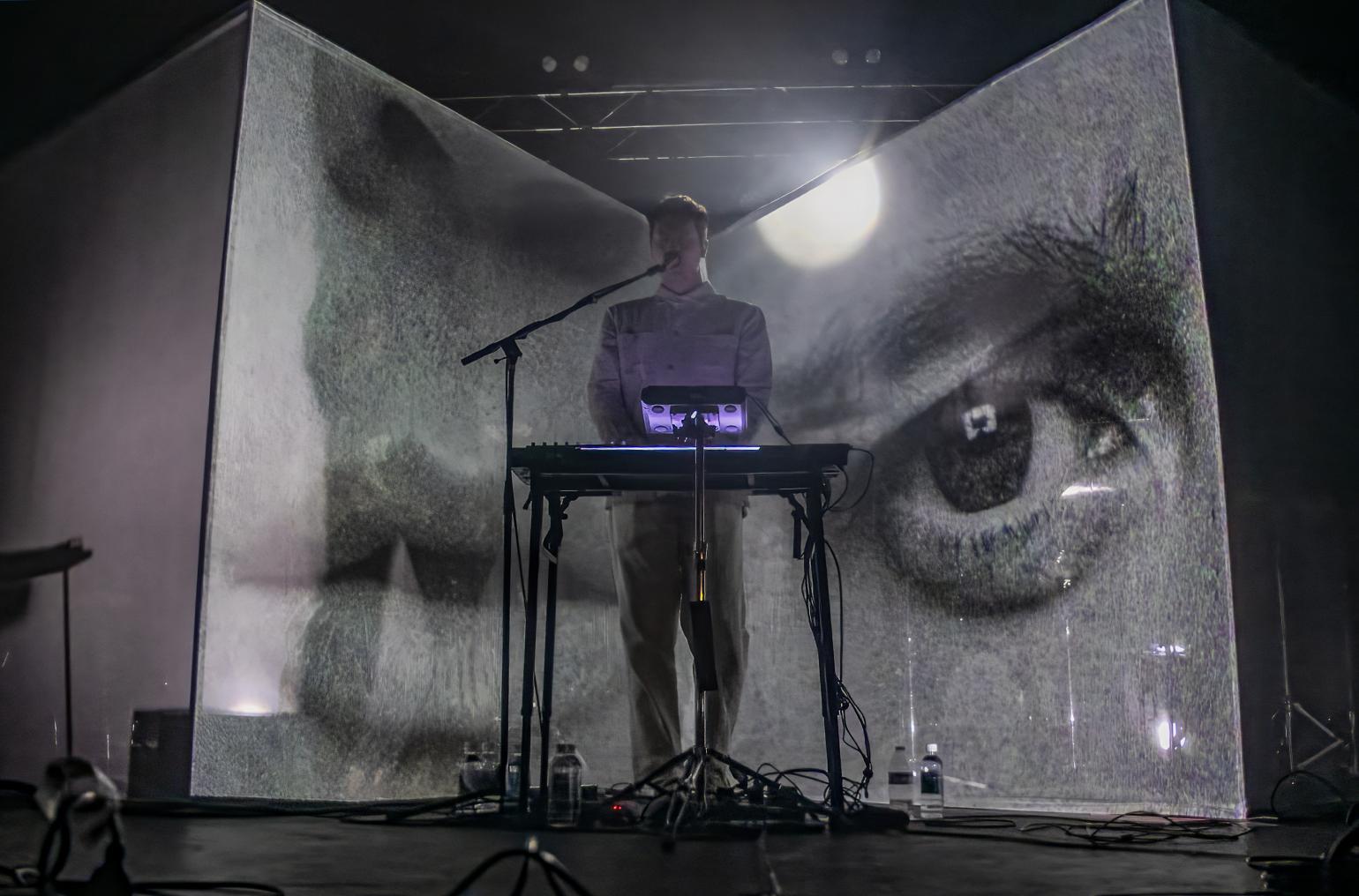 Musician playing experimental music on stage against the backdrop of a large human eye. Photo by Tom Beck