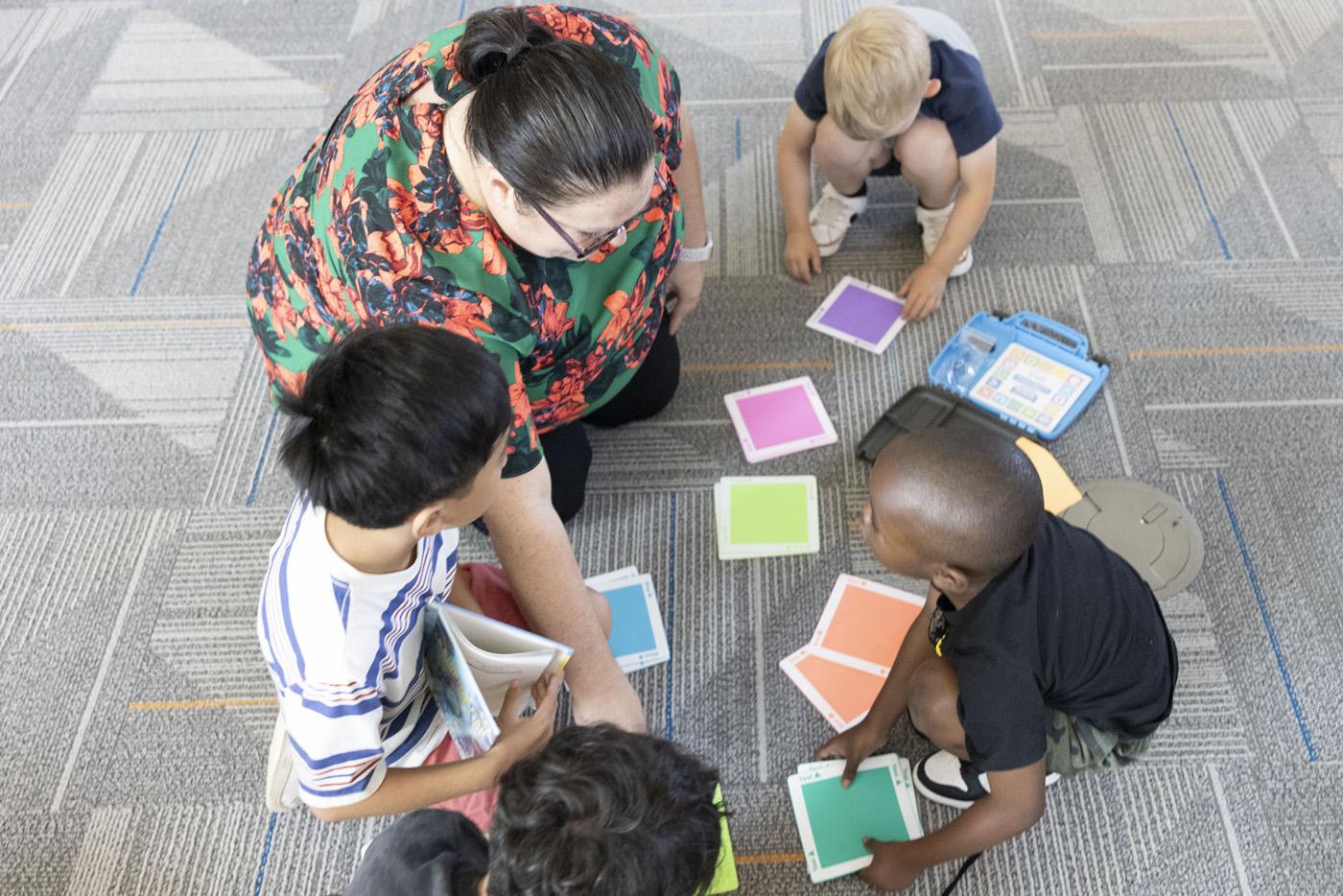 A teacher sits on the floor with students using color squares for lesson.
