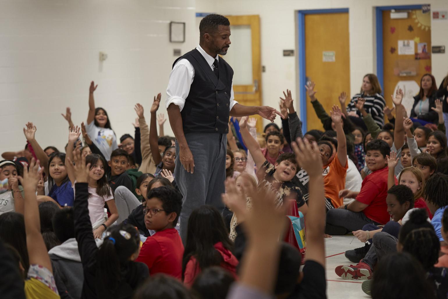 Actor and teaching artist Mikey Wiley surrounded by students in a classroom engaging with his teachings with their hands raised 