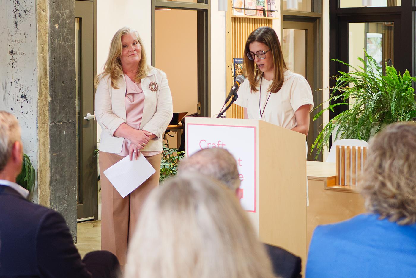 A woman speaks at a podium while another woman looks on.