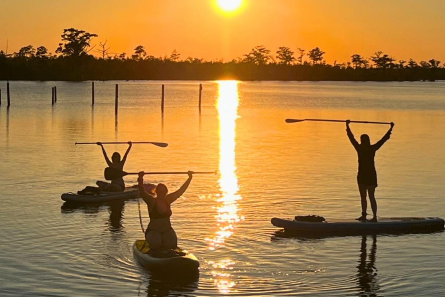 Three kayakers on the water with a sunset behind them.