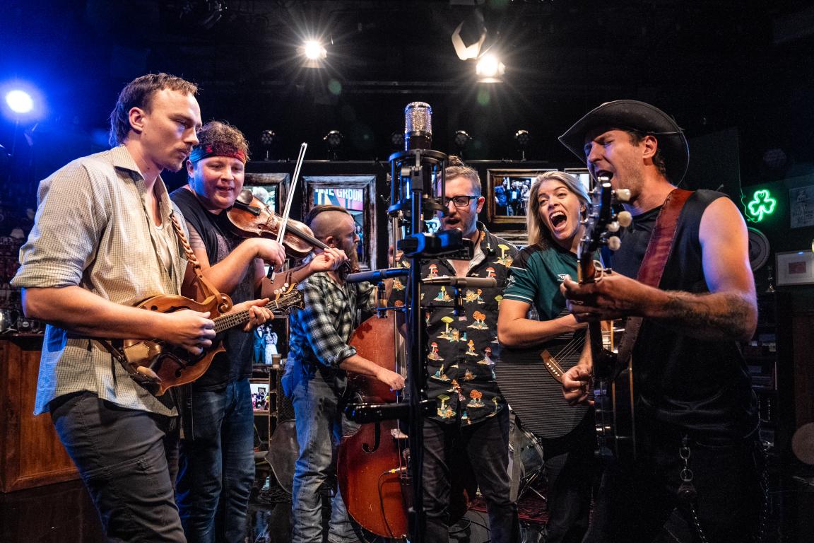 Six members of The Tan & Sober Gentlemen sing and play around one microphone. 