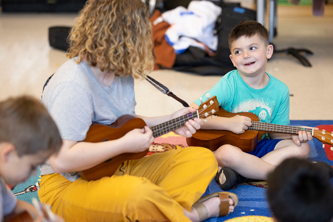 teacher sitting on the ground with young students around her, everyone is playing guitar