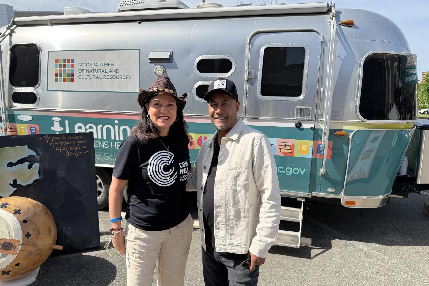 NC DNCR Secretary Pamela Cashwell wearing a Come Hear NC logo t-shirt, standing in front of of a DNCR branded airstream next to Al Strong, a 2x Grammy Nominated trumpet player, composer, recording artist and educator