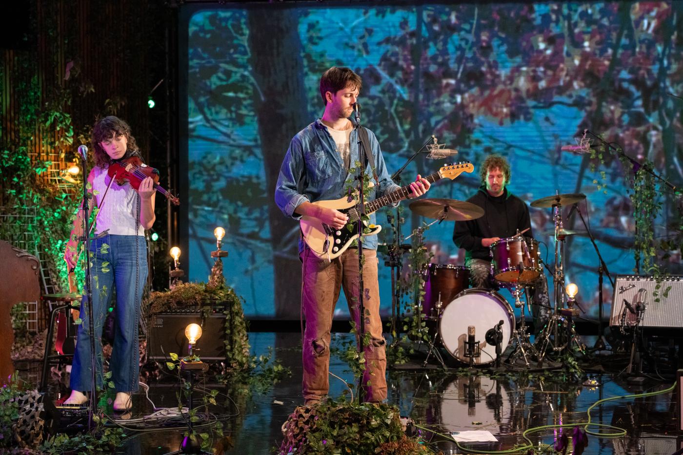 Sluice's fiddler, lead singer and drummer play on stage with multimedia scenes of nature in the background