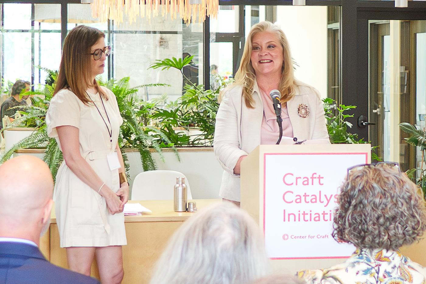 A woman standing at a podium speaking with another woman standing nearby.
