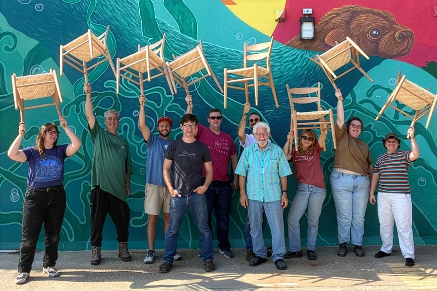 A group of people holding chairs above their heads in front of a mural.