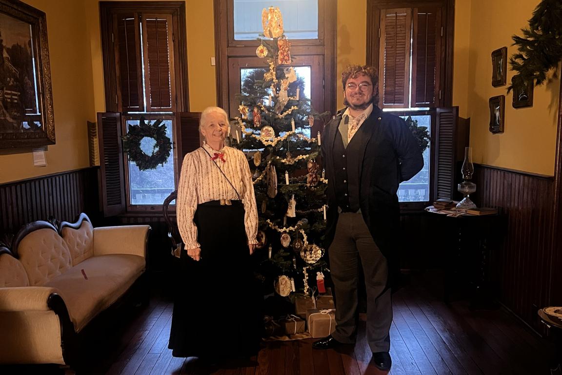 Woman and a man dressed in Victorian clothes in front of a Victorian Christmas tree.