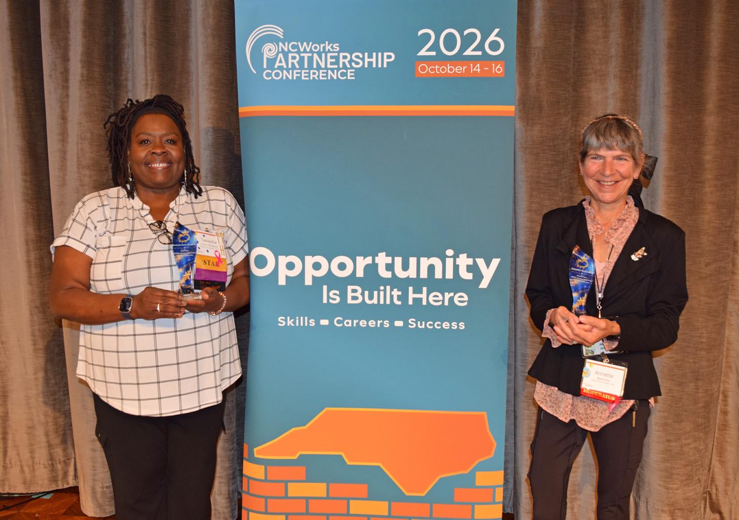 Two women holding their award plaques, with a sign between them that says "Opportunity is Built Here: Skills, Careers, Success"