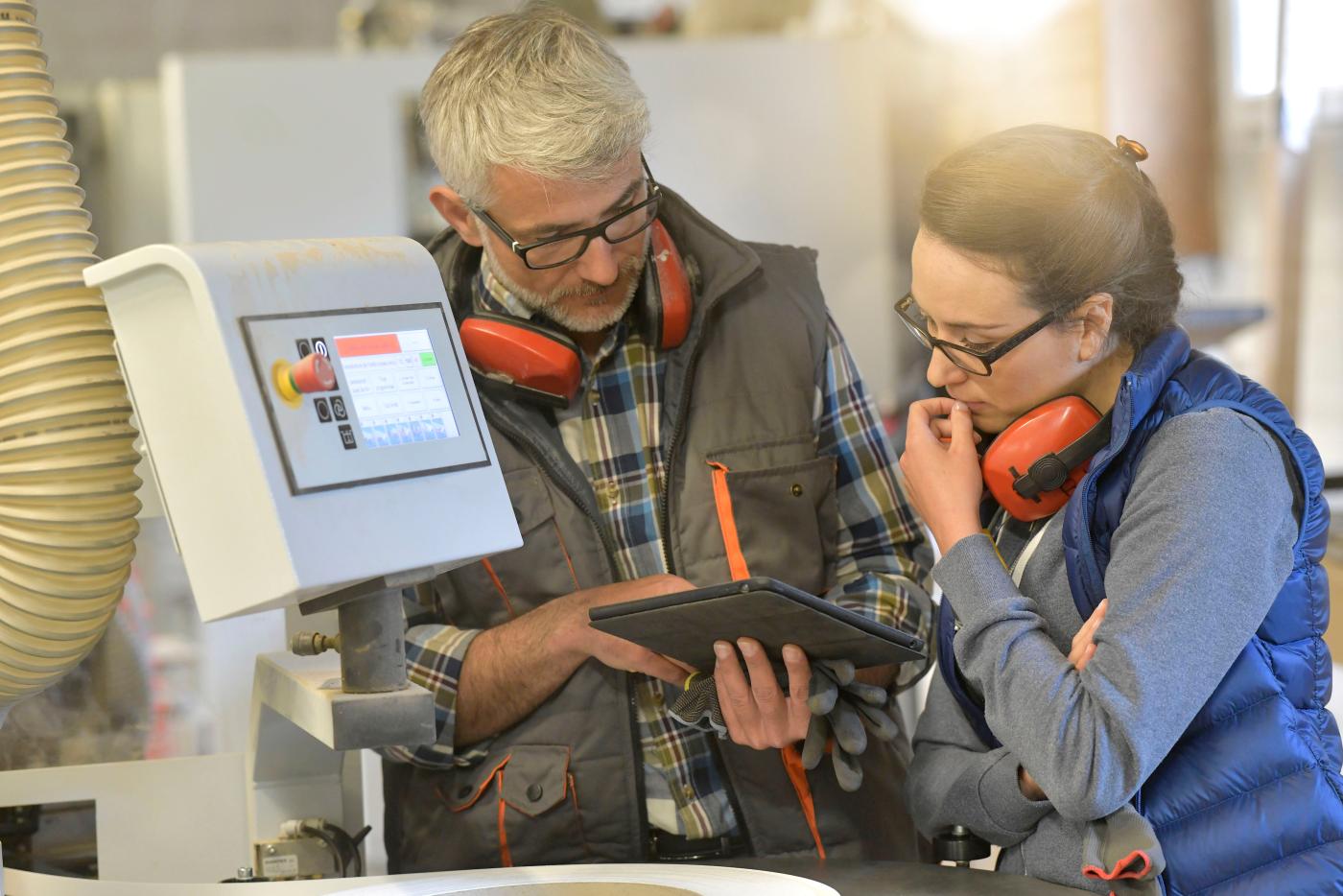 Two workers wearing hearing protection and glasses review information on a tablet next to industrial machinery in a manufacturing facility.