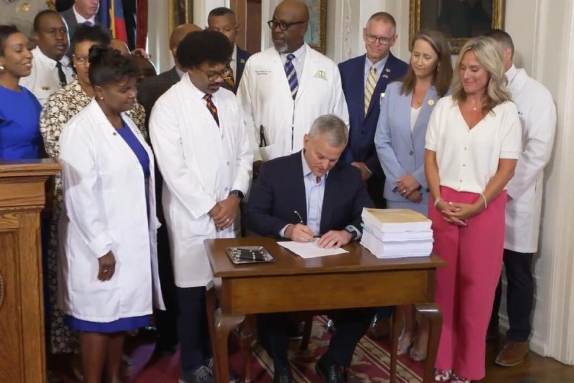 Governor signs paper while at desk surrounded by many people