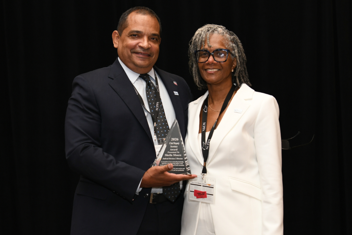 Smiling man in a dark suit and woman in a white suit proudly pose with a glass award against a black backdrop at a formal event.