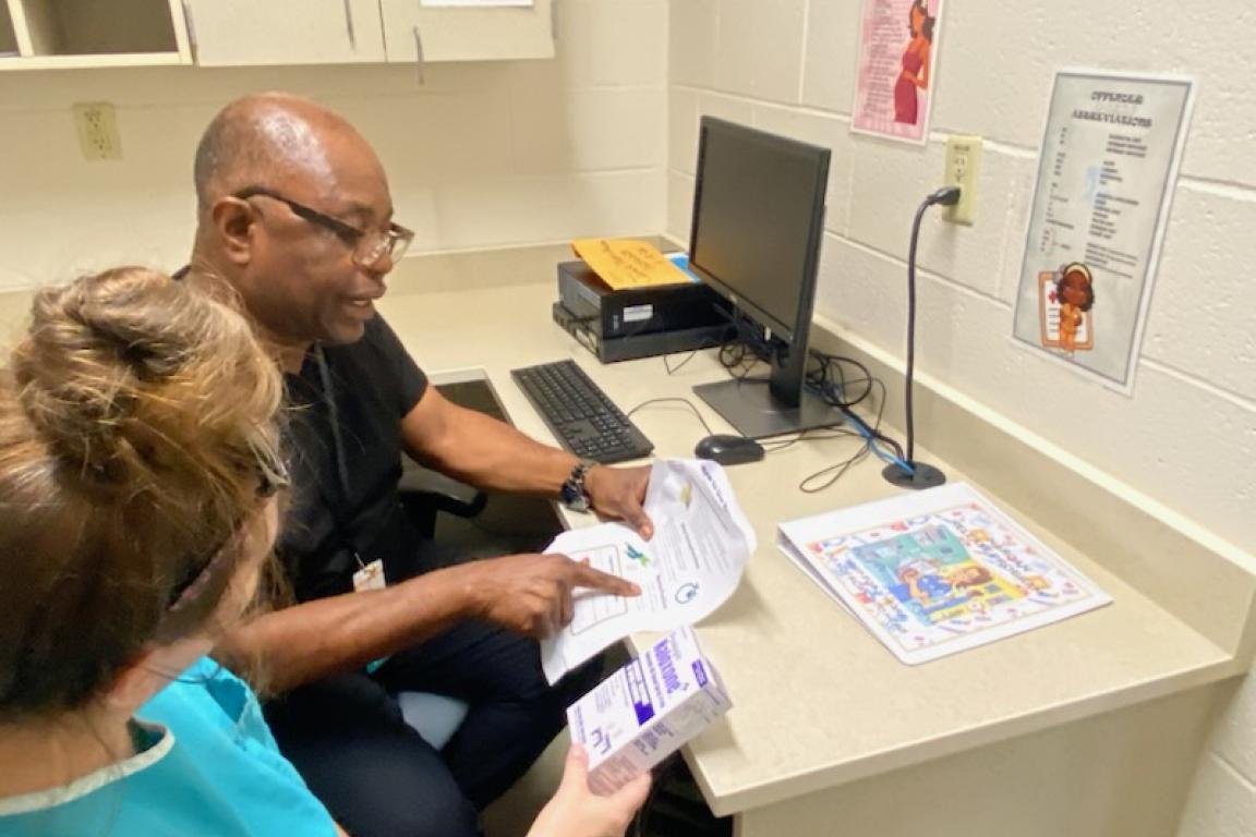 A man and a woman review documents and medication in an office with a computer, posters on the wall, and educational materials on the desk.