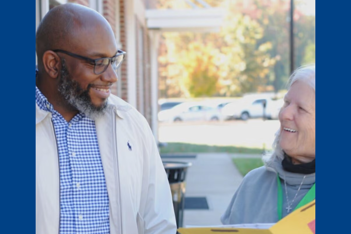 A smiling Black man and white-haired woman chat outside a building