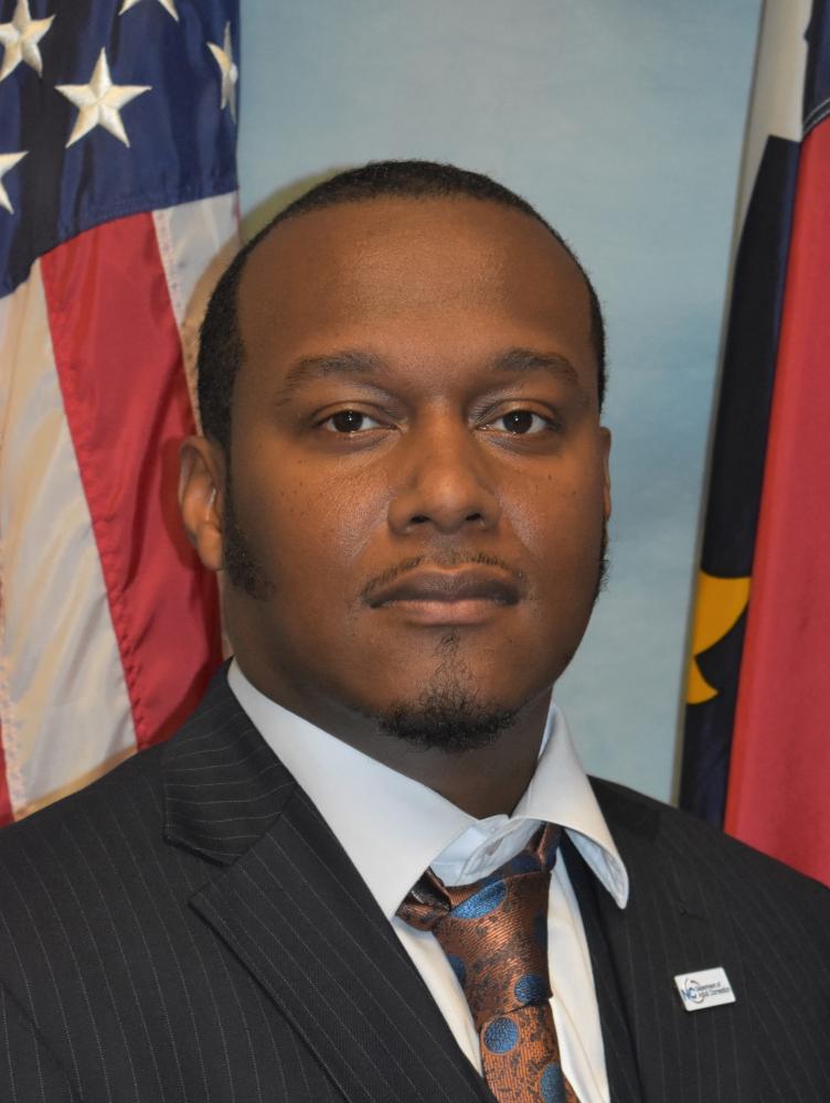 Linwood Etheridge in jacket and tie between flags of United States and North Carolina