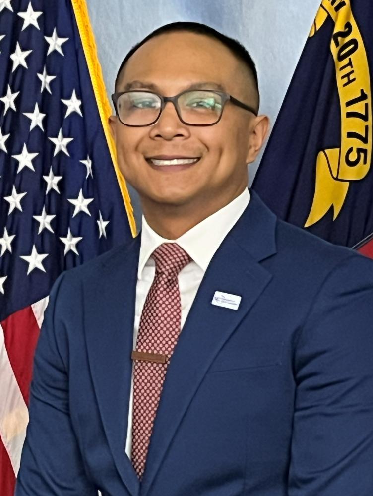 Smiling man in glasses and a blue suit with a red tie stands in front of American and state flags.