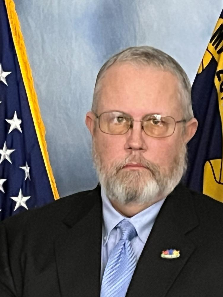 A man in a suit with glasses and a beard stands in front of the North Carolina and American flags, conveying a formal and solemn tone.