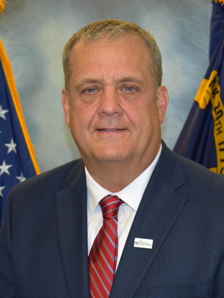 Formal headshot of a middle-aged man in a dark suit and red striped tie, posed before U.S. and West Virginia flags