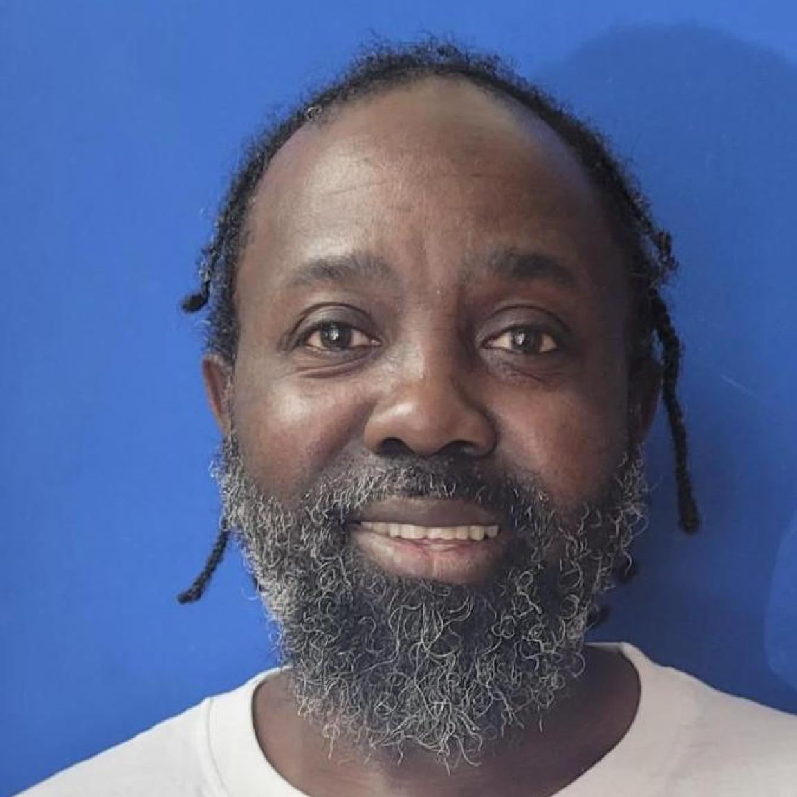 Smiling man with a gray beard and short dreads against a blue background, wearing a white shirt. 