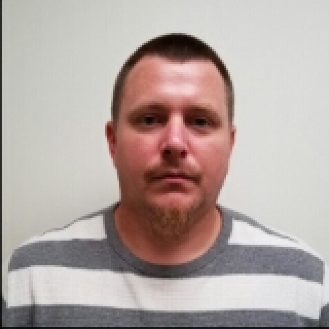 A man with short hair and a goatee wearing a striped shirt stands against a plain white background. 