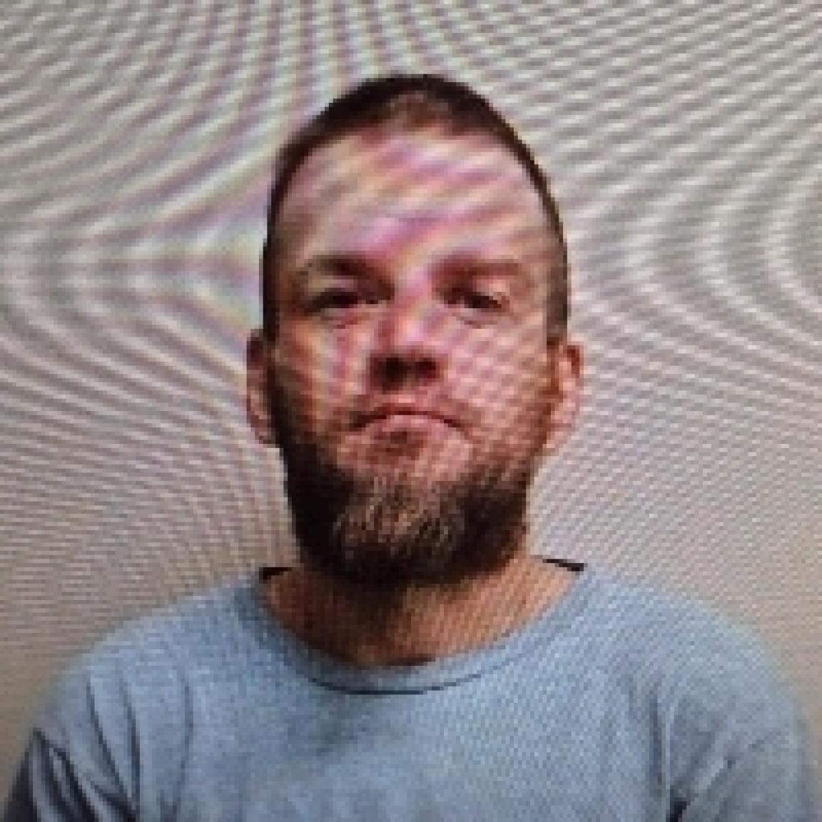 A bearded man in a gray shirt is sitting against a textured, patterned wall. 