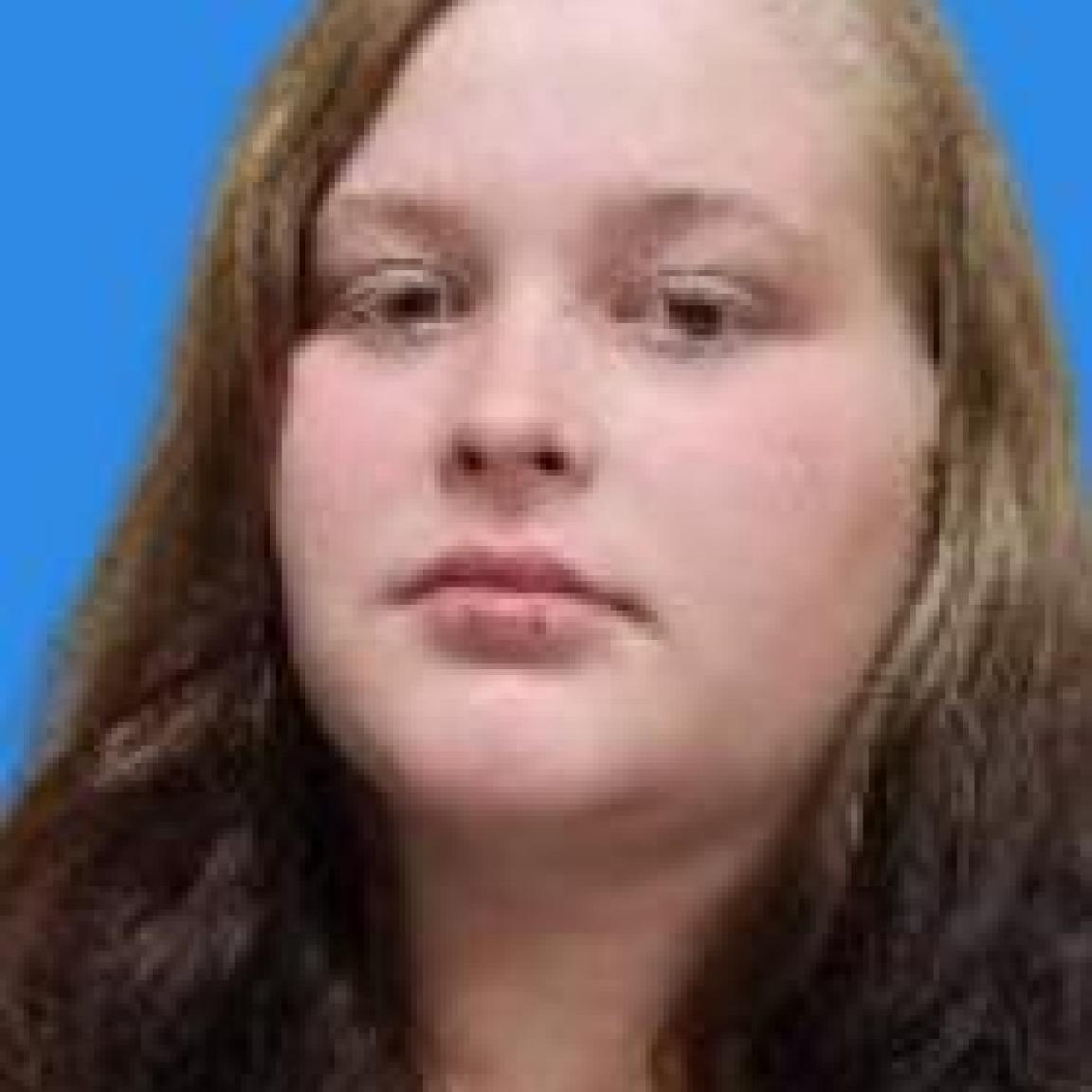 A woman with long, wavy brown hair is set against a plain blue background. Her expression is neutral