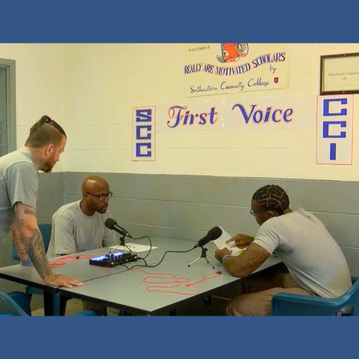 Three men in a prison setting are engaged in a podcast recording. Two sit at a table with microphones, while one stands beside them, appearing attentive and focused. A sign above reads "First Voice" with SCC and CCI logos. 