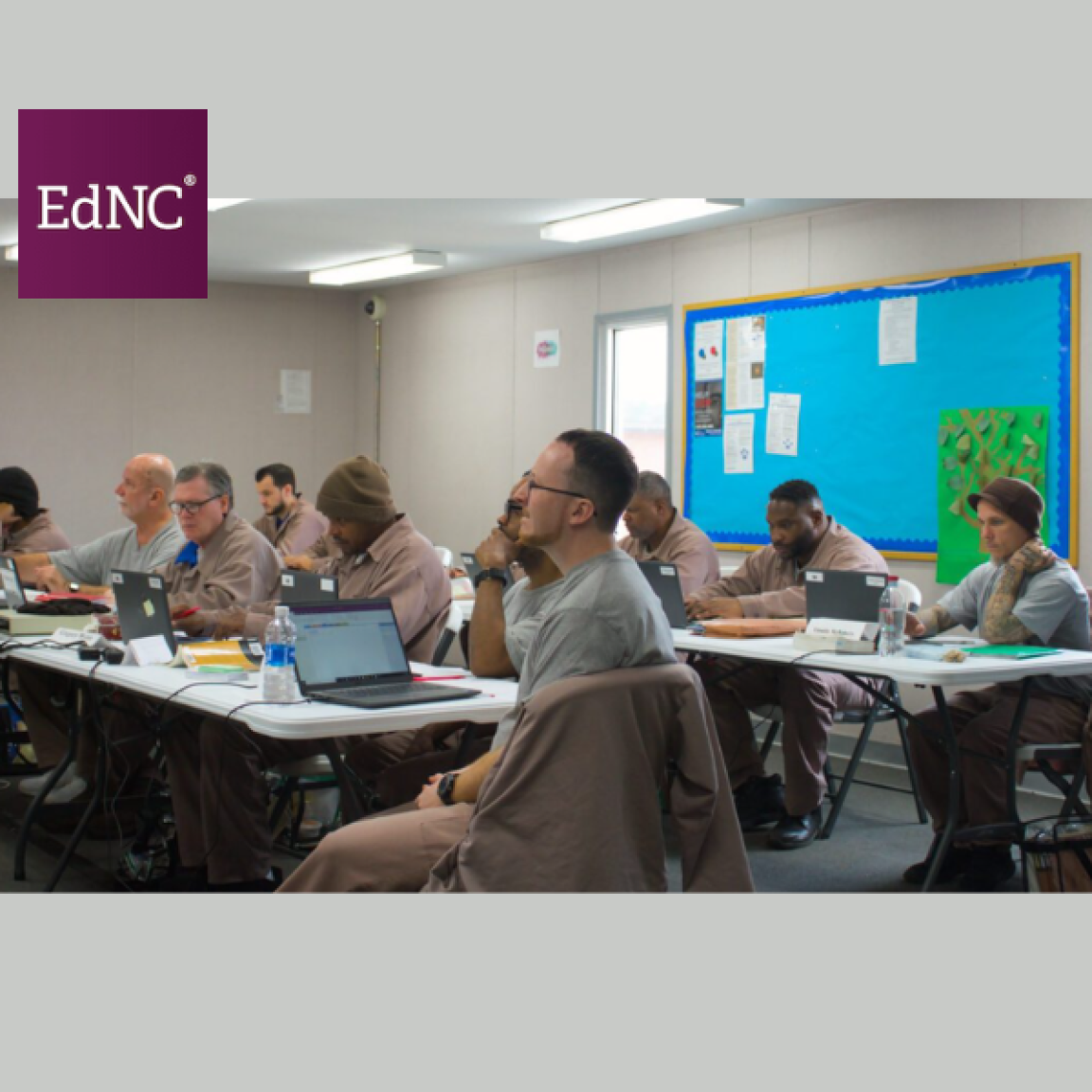 A group of men in uniforms sit at tables in a classroom, engaging with laptops. The room has white walls, a bulletin board, and fluorescent lighting.