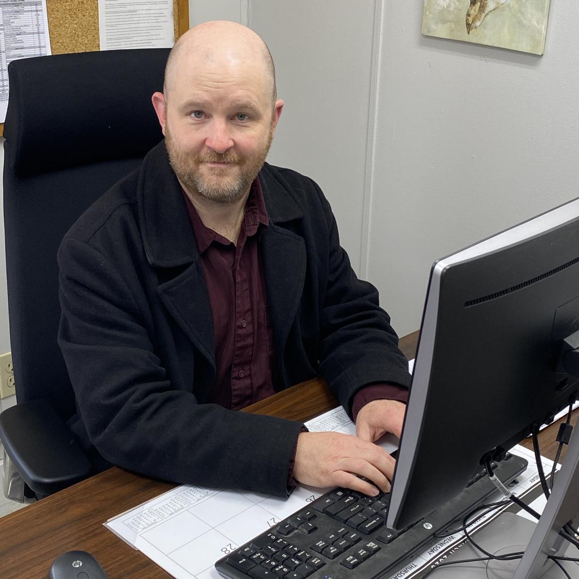NCDAC social worker Ashley Penny sits in front of his computer. He is wearing a dark jacket and maroon shirt.