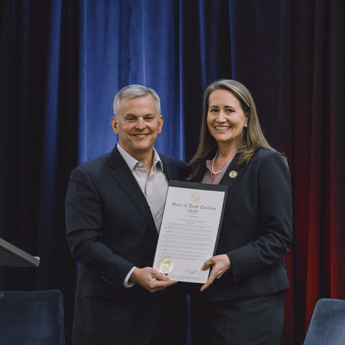 A man and a woman stand smiling, holding a certificate reading "State of North Carolina" on stage.