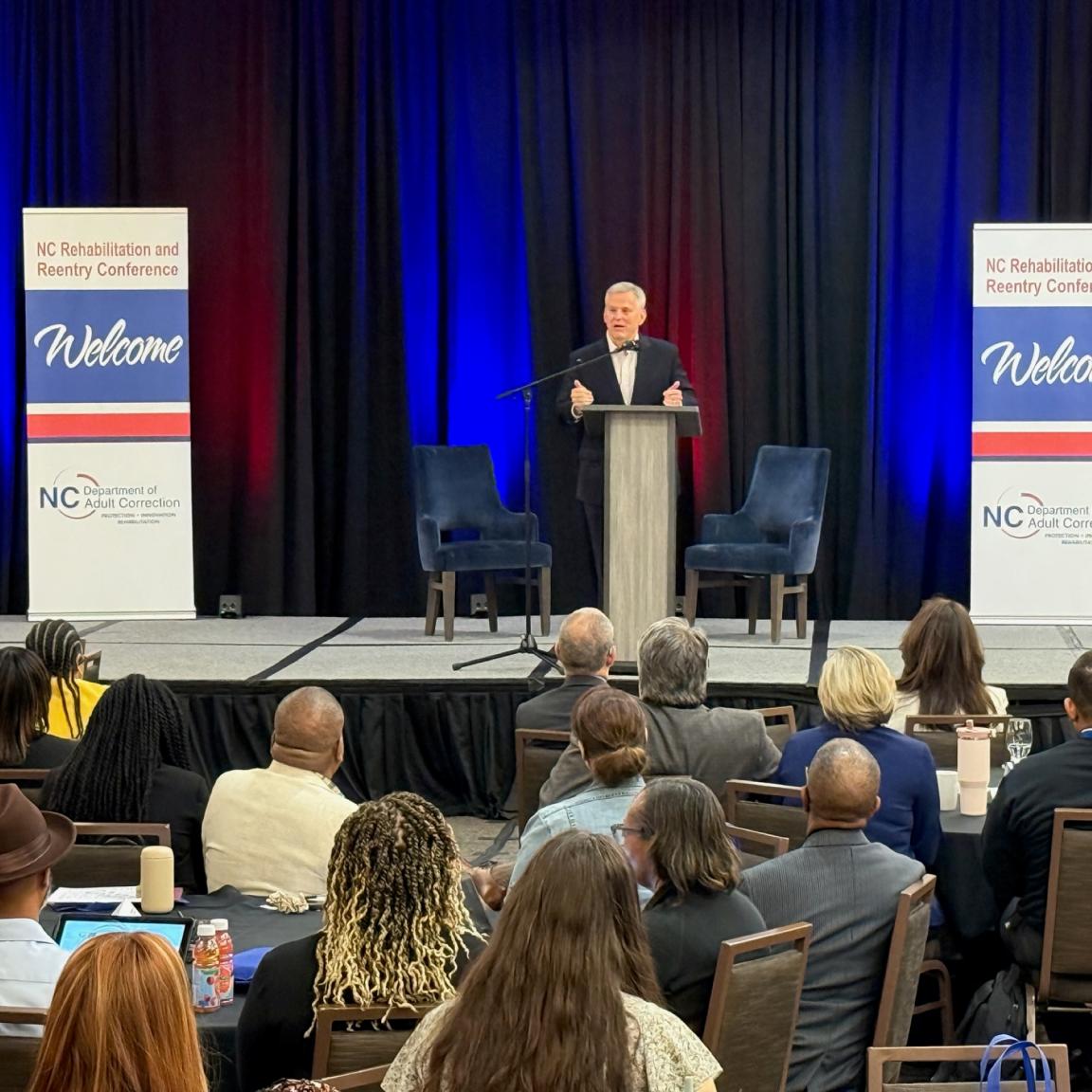 Gov Josh Stein stands at a podium on stage at the NC Rehabilitation and Reentry Conference. Attendees are seated, listening intently. 