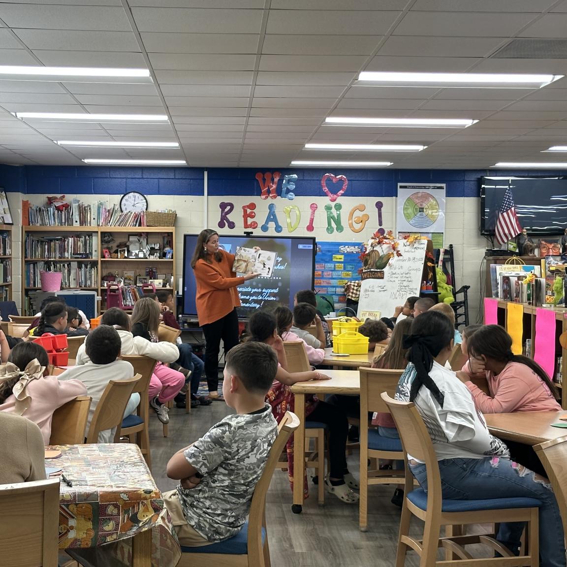 a woman stands at front of classroom reading book to kids