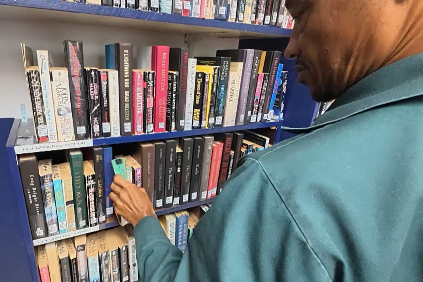 A man in a teal jacket browses colorful books on a library shelf, reaching toward a title.