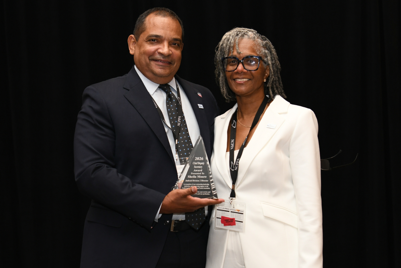 Smiling man in a dark suit and woman in a white suit proudly pose with a glass award against a black backdrop at a formal event.