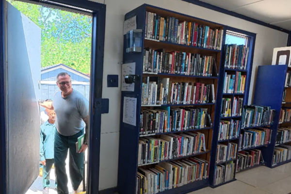 A smiling man stands in a library doorway holding a green book, with another person behind him and tall shelves packed with books.