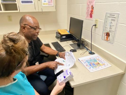 A man and a woman review documents and medication in an office with a computer, posters on the wall, and educational materials on the desk.