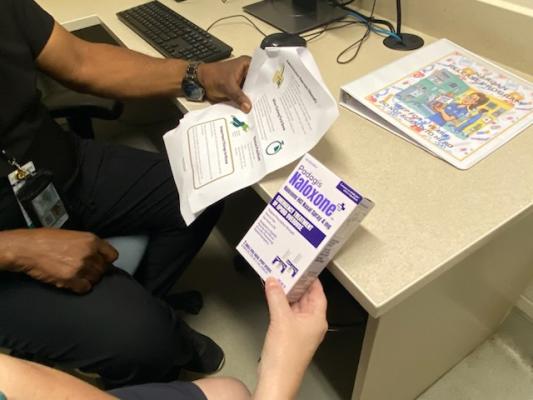 A seated person in a black uniform hands over a folded brochure beside a colorful map on a desk, suggesting a calm, helpful information exchange.