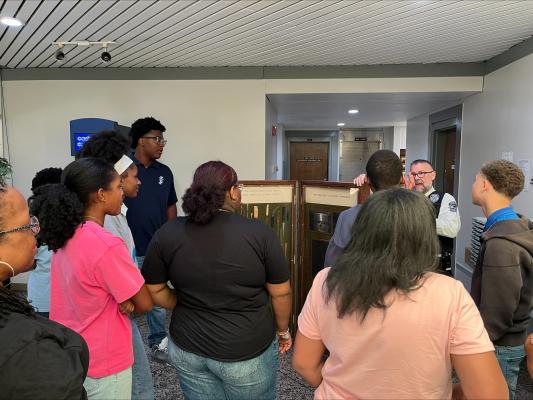 Teens and adult chaperones listen to a Central Prison staff member explain displays about contraband and prison security during an educational tour.