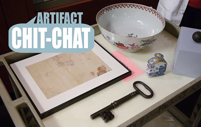 A promotional image titled "ARTIFACT CHIT-CHAT" features a group of historical artifacts on a display tray. Items include a framed antique map with light staining, a large porcelain punch bowl decorated with various motifs, a small covered blue and white porcelain container, and a large, old iron key.