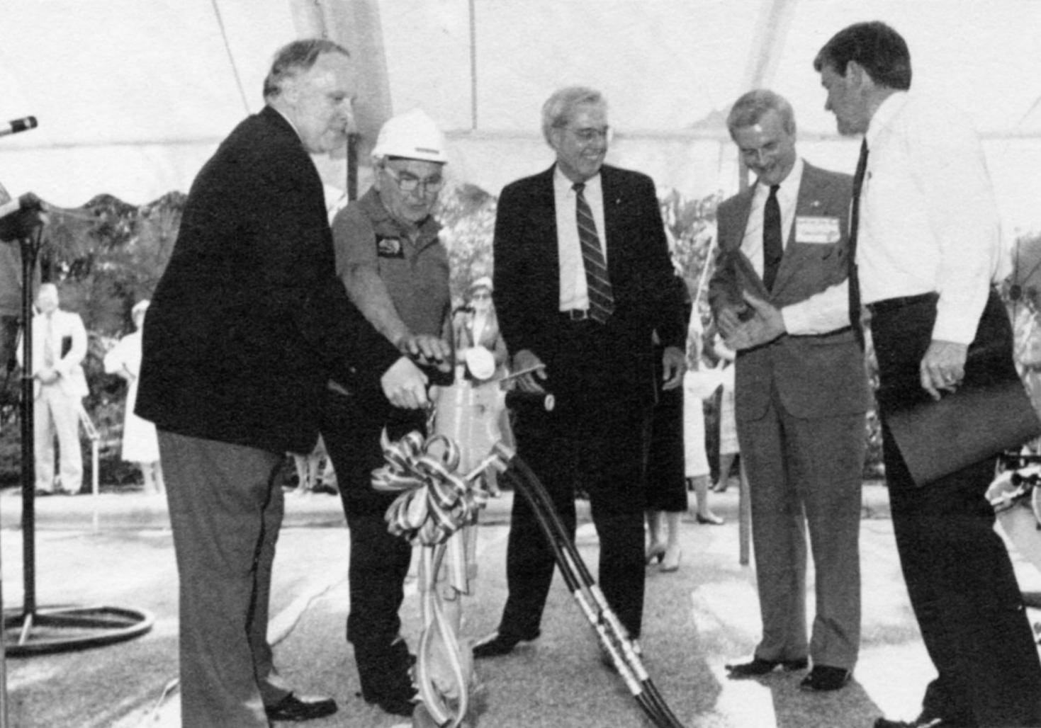 Black-and-white photograph of several men in suits and a construction worker gathered under a tent, participating in a ceremonial groundbreaking using a drill adorned with a ribbon.