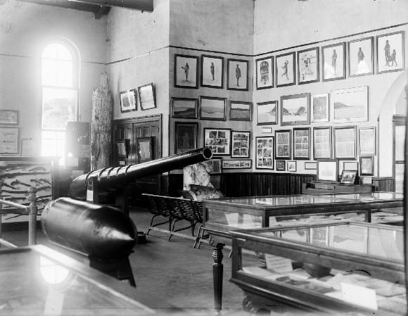 Black-and-white image of a historic museum gallery featuring a large cannon, glass display cases, and walls lined with framed images and documents.