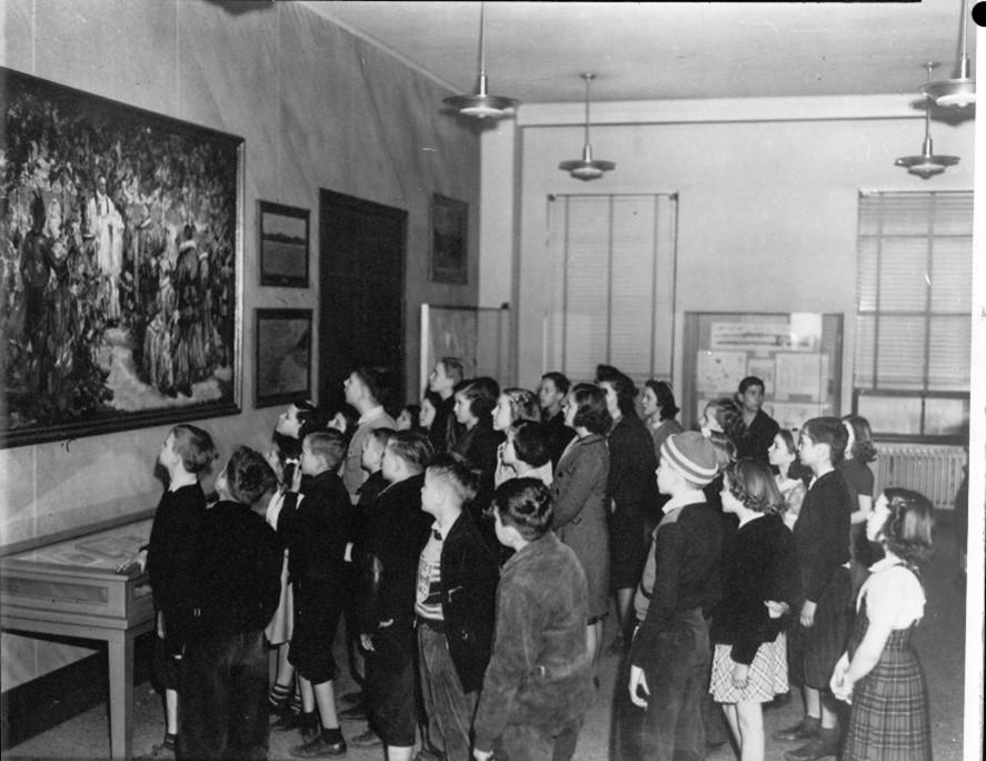 Black-and-white photograph of a group of schoolchildren gathered in a museum gallery, viewing a large painting on the wall.
