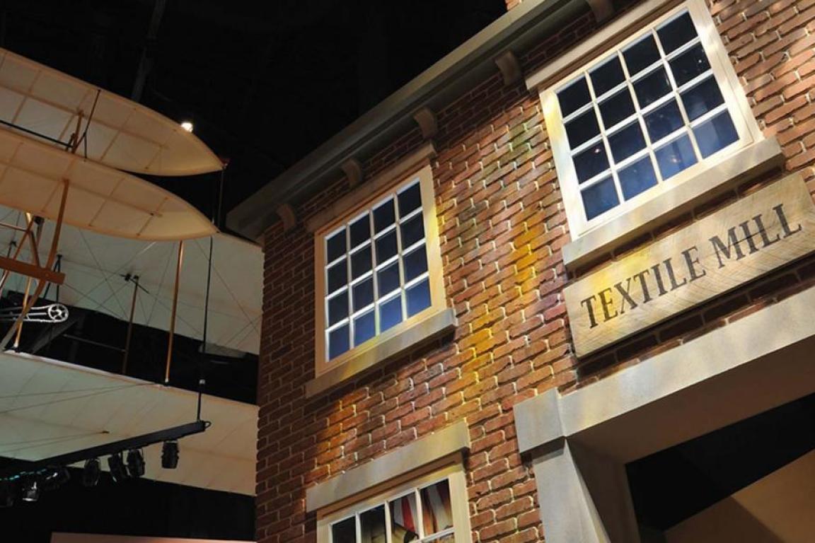 A museum display featuring a brick facade of a "TEXTILE MILL" with large, multi-paned windows. An early biplane glider model hangs from the dark ceiling in the background.