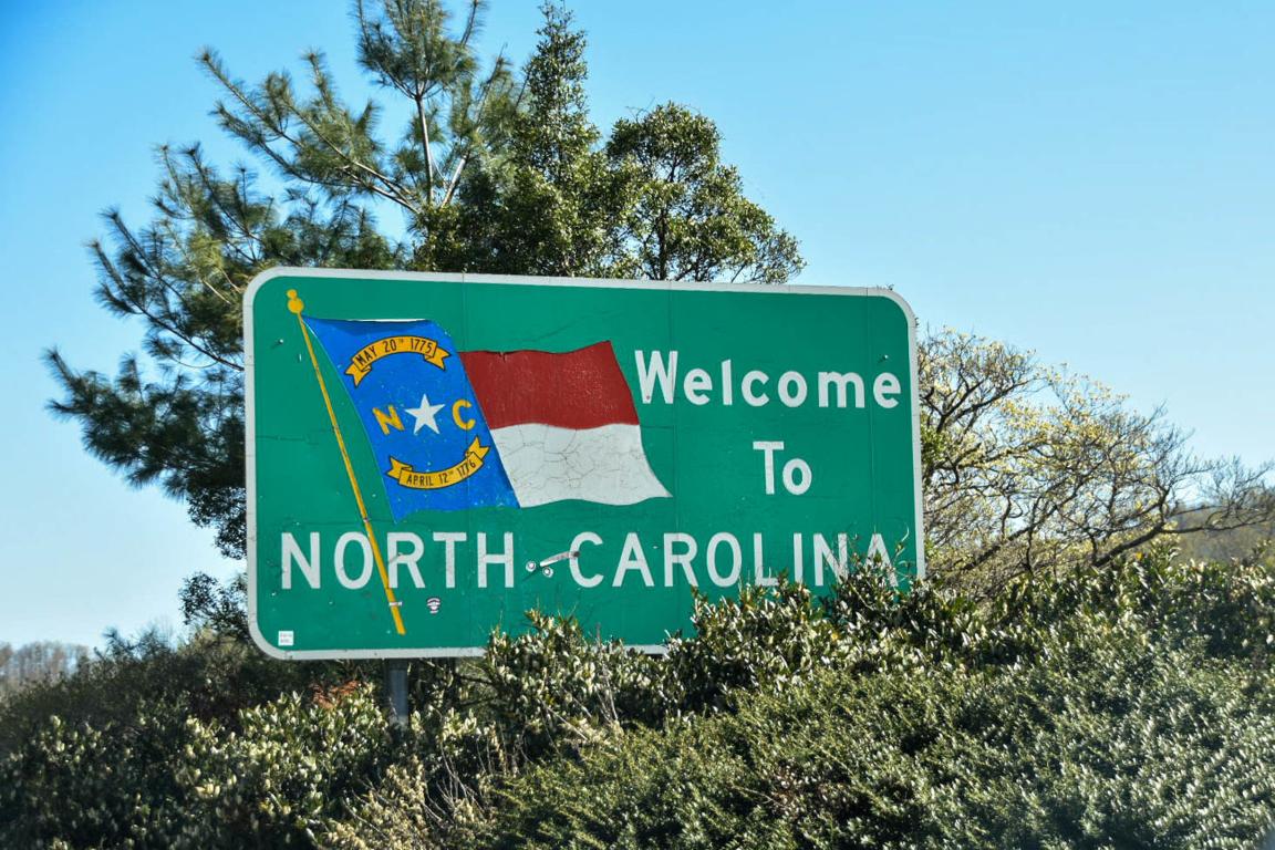 A green sign with the NC flag that says Welcome to North Carolina. The sign is nestled in green trees with a blue sky background. 