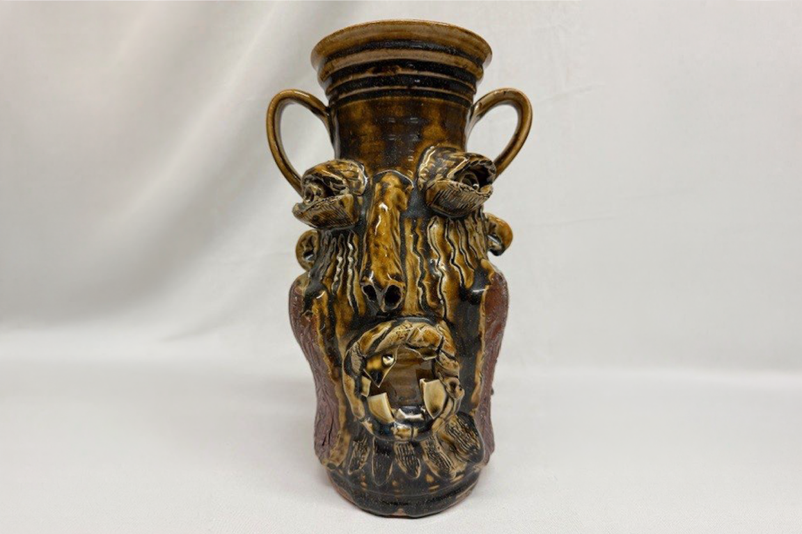 An earthenware face jug with a shiny brown glaze and two small handles at the top. The jug features an exaggerated, sculpted face with bulging eyes, a long nose, and a wide-open mouth showing several jagged white teeth.