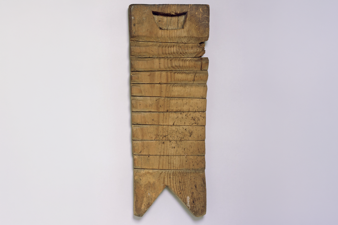 A vertical photograph of a wooden washboard shows a single plank of deeply grooved wood, with a roughly carved, shallow, boat-shaped indentation at the top. The sides are rugged and a split is in the upper right side.