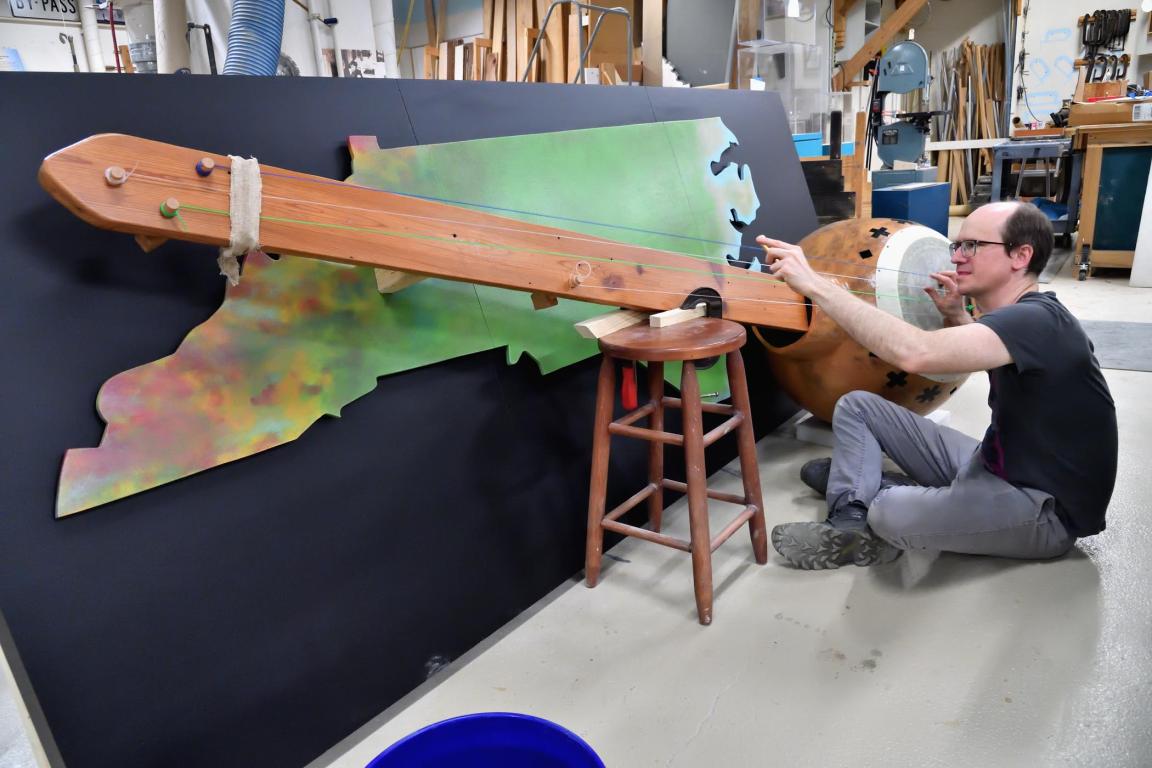 Man adjusts strings on a large, handmade banjo mounted on a North Carolina-shaped backdrop in a workshop.