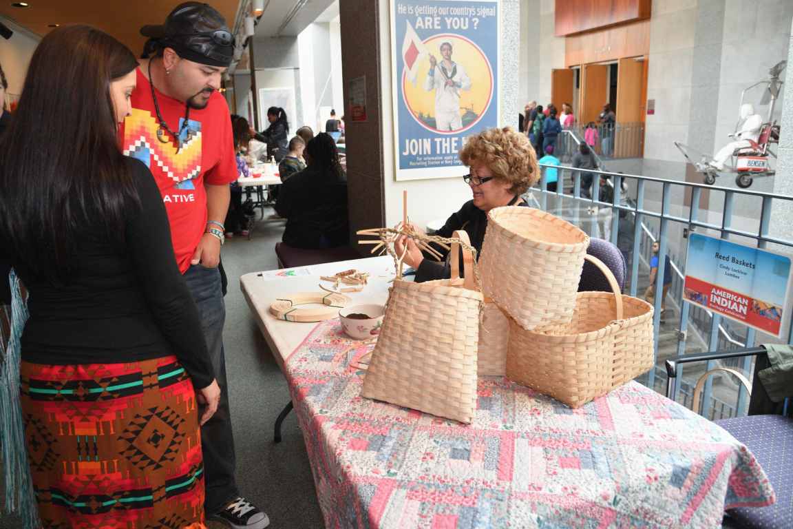 A woman demonstrates basket weaving at a table with finished reed baskets while two visitors watch.