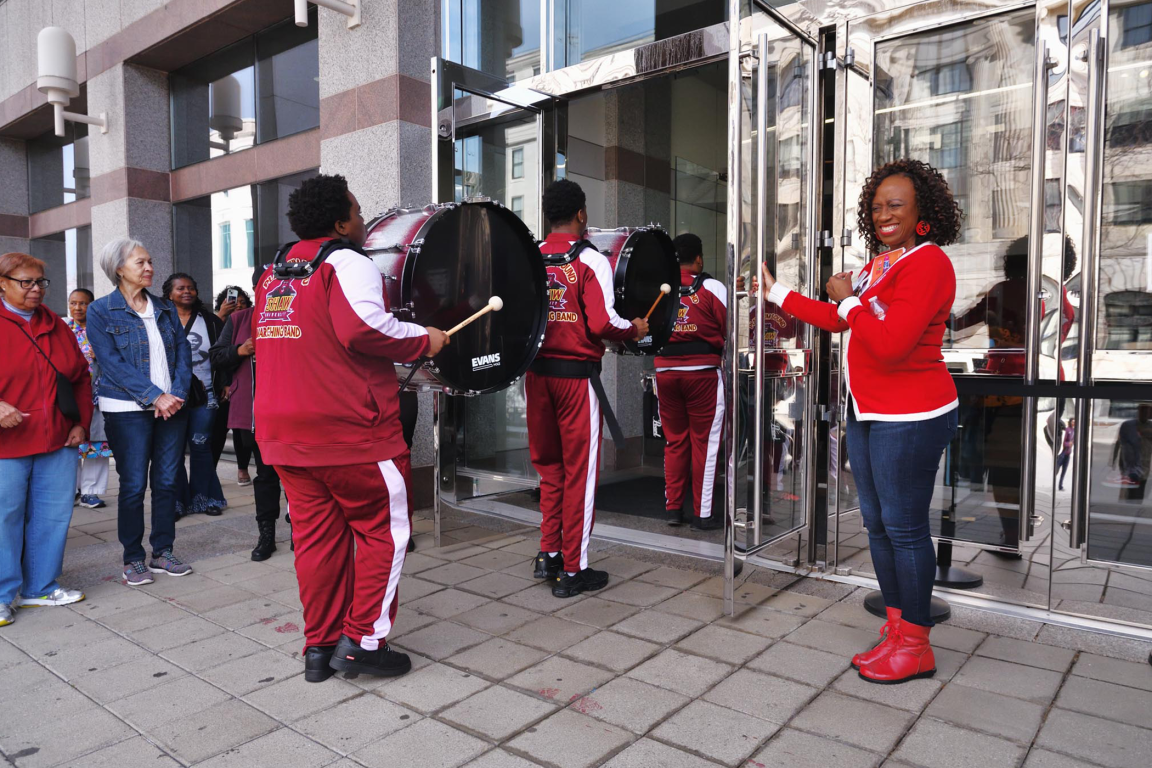 A smiling woman in a red cardigan holds open a glass door as members of a marching band carrying bass drums enter a building, while spectators watch from the sidewalk.